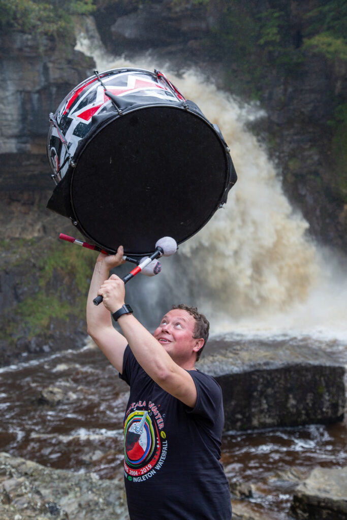 A drummer olding a bass drum in the air in front of a waterfall