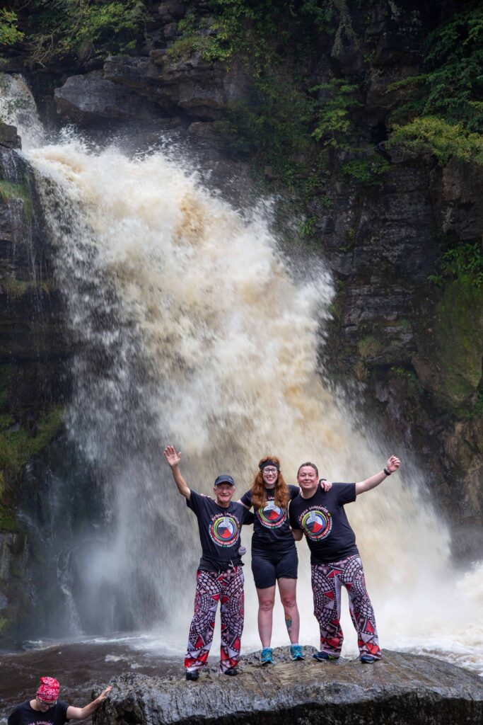 3 memebers of Batala standing on a rock in front of the waterfall