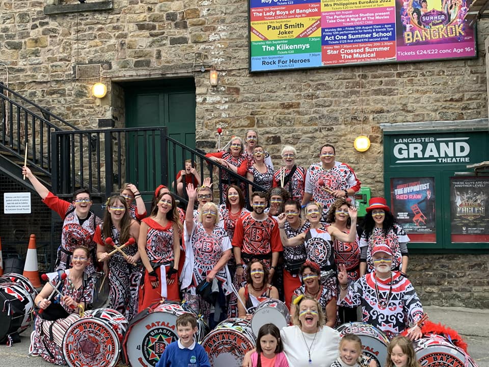 Batala lancaster grouped together at the side of the Lancaster Grand Theatre, wearing their red, black and white costumes